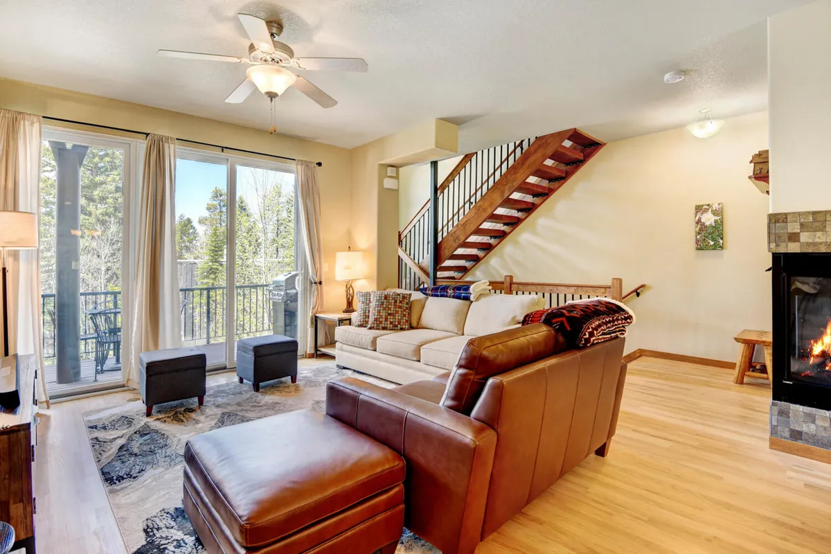 A sunlit, open-concept living area with a plush sofa facing a unique gas fireplace. Large windows offer views of tranquil mountain landscapes, while the seating arrangement encourages both relaxation and group conversation. The ambiance is cozy, enhanced by the natural light and welcoming fire.