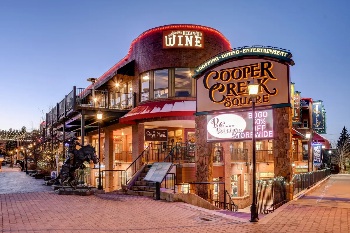 A lively exterior shot of Cooper Creek Square, featuring its inviting sign, a wine shop, and twinkling decorative lights, surrounded by cheerful activity and festive ambiance.