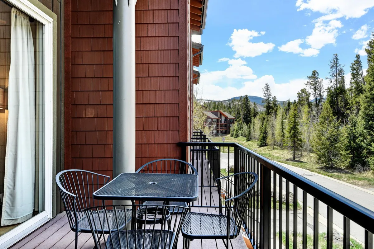 An inviting balcony or patio space with a table and chairs, natural wood siding, and a scenic mountain view in the background. The setting is serene, alluding to peaceful mornings or evenings enjoying the outdoors.
