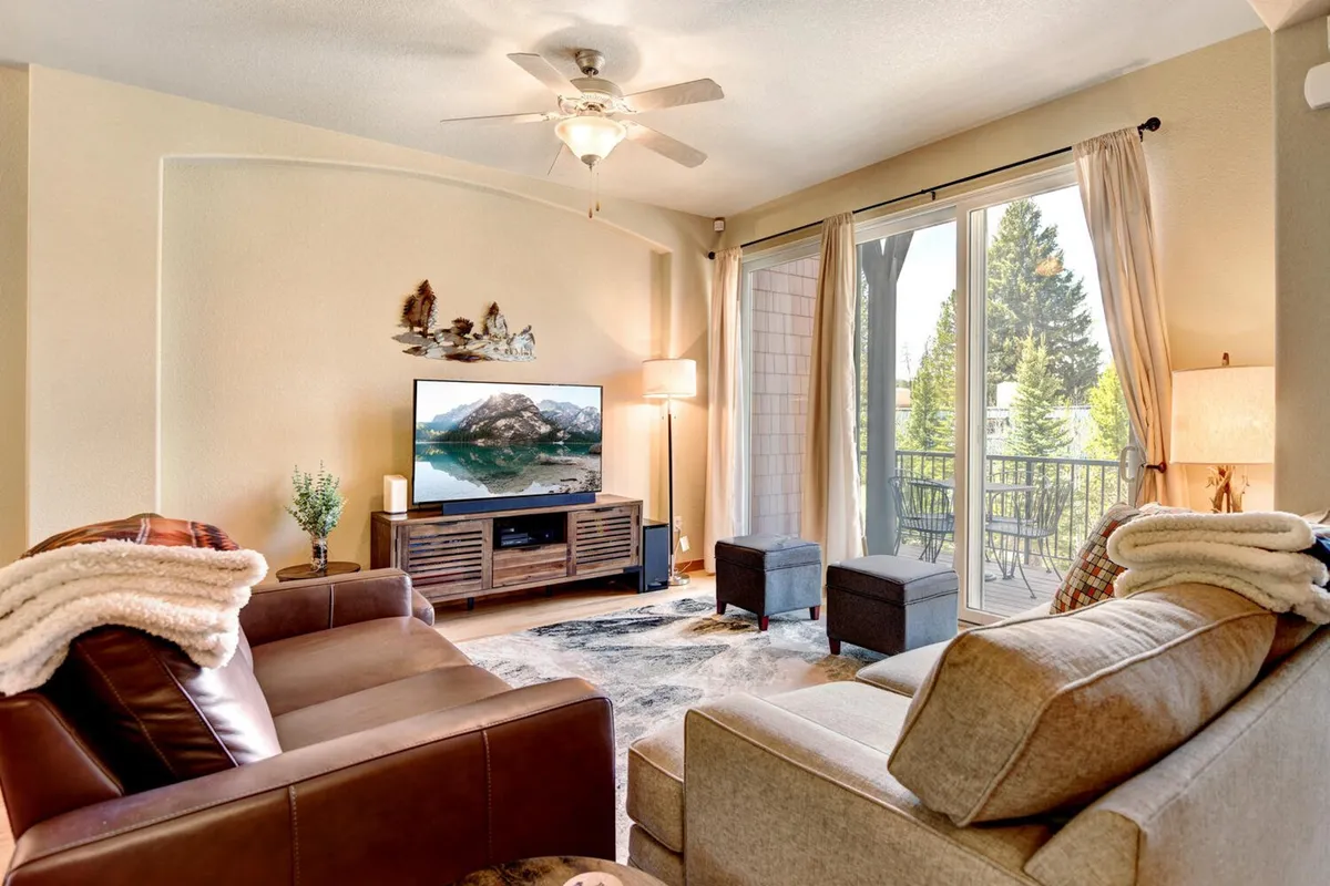 A comfortable living area featuring a spacious sofa, large rug, and a wall-mounted HD smart TV. Expansive windows fill the room with natural light and frame scenic views. The space feels ideal for both cozy movie nights and family gatherings.