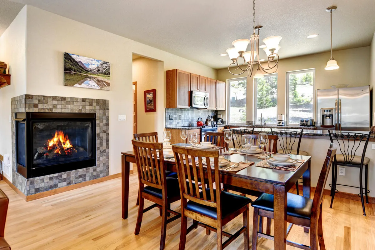 An open dining area centered around a large table, with a cozy fireplace, kitchen island, and expansive windows. The setup invites group meals, relaxed gatherings, and lingering over coffee or games, embodying a social dining nook.