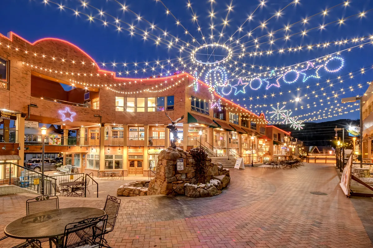 This exterior image features a lively outdoor plaza illuminated by overhead string lights, decorative star motifs, a stone water feature, and inviting outdoor seating. The area is arranged for social gatherings and holiday festivities, with an atmosphere that suggests community events, outdoor dining, and evening entertainment.