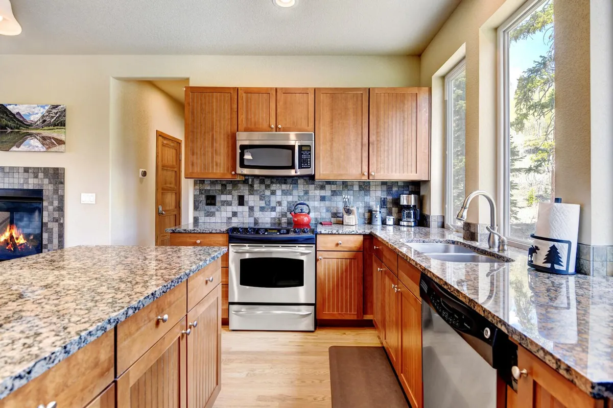 A spacious, well-lit kitchen featuring granite countertops, stainless steel appliances, a fireplace, and large windows filling the space with natural light. The layout is ideal for cooking and entertaining, with visible tools and ingredients suggesting a culinary space ready for group meals.