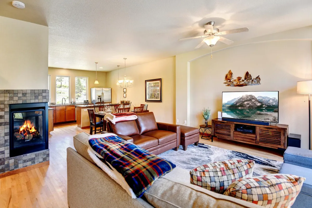 A welcoming open-plan living and dining space with a plush sofa, glowing fireplace, dining area, and a large TV positioned near a window with a mountain view. The space is arranged for both family gatherings and relaxing by the fire.