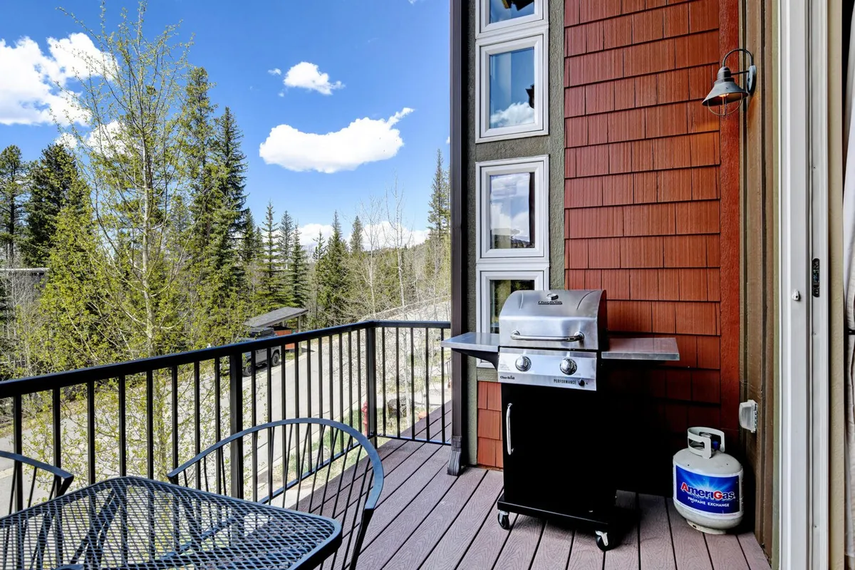 A private patio area featuring a gas grill, comfortable outdoor seating, and panoramic views of trees and distant mountains. The scene suggests an inviting space for barbecuing, relaxing, or enjoying mountain air after a day of adventure.